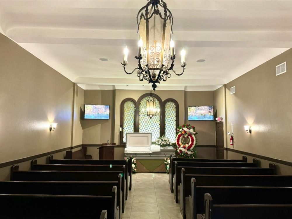 Interior of a funeral chapel with dark pews, casket, and floral wreath; chandelier hangs from ceiling.