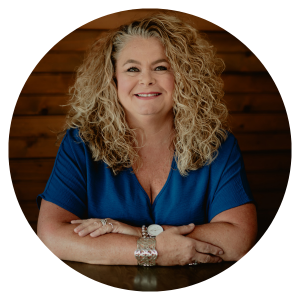 Smiling woman in a blue blouse with curly blonde hair, leaning on a table against a wooden wall.
