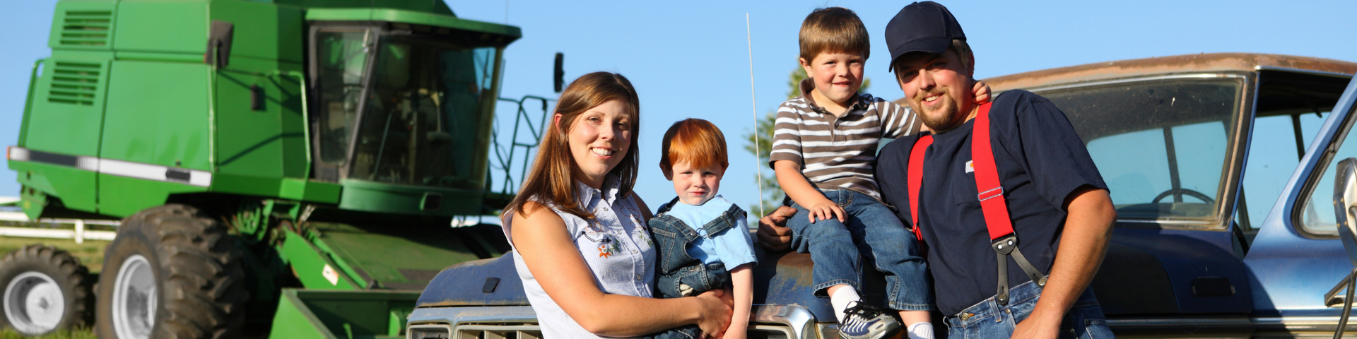 Family standing by green farm equipment and a blue tractor, smiling outdoors.