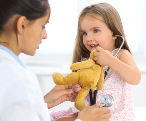 Doctor examining a smiling child holding a teddy bear in a clinic