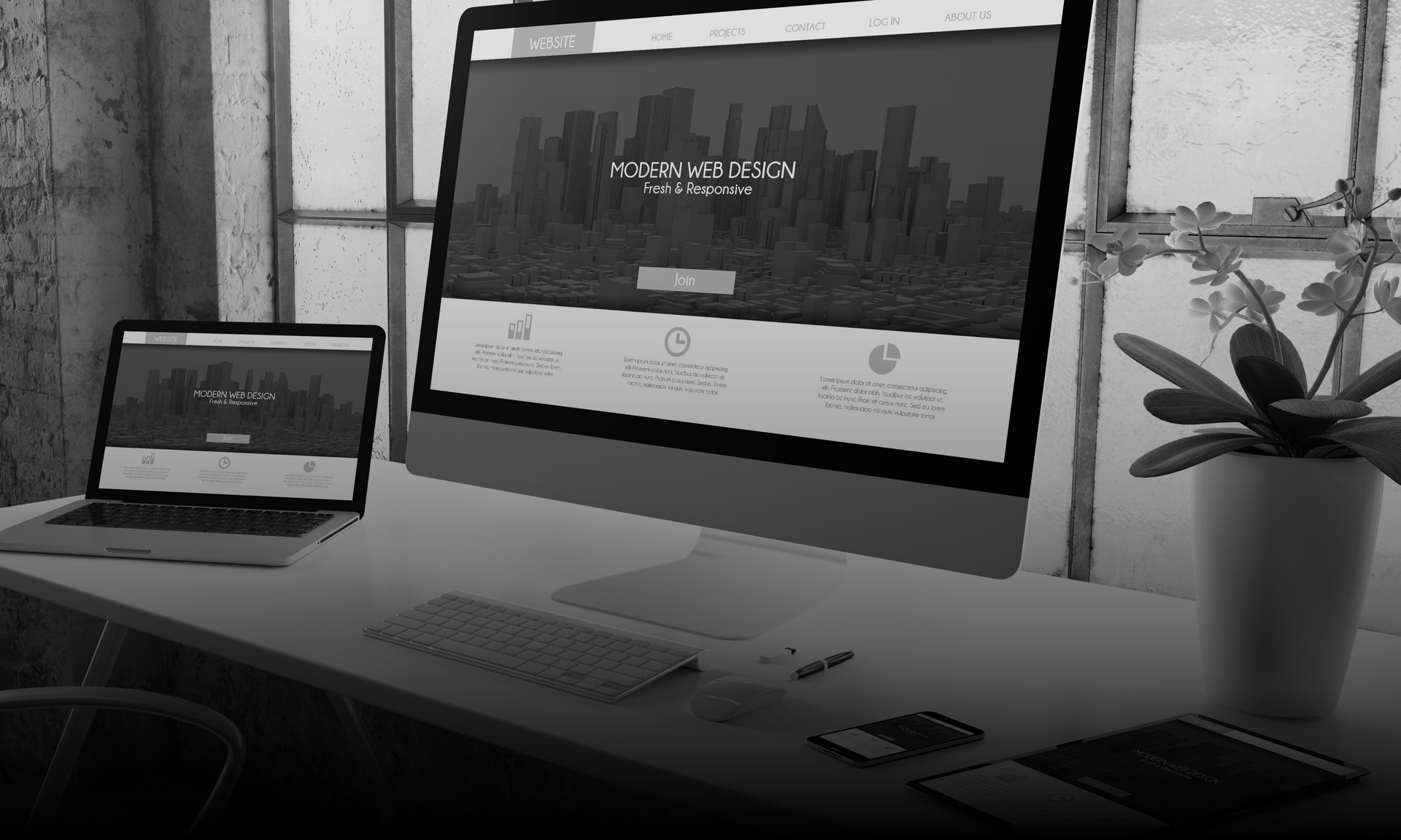 A laptop and desktop computer displaying a website with city skyline, on a desk by a window.