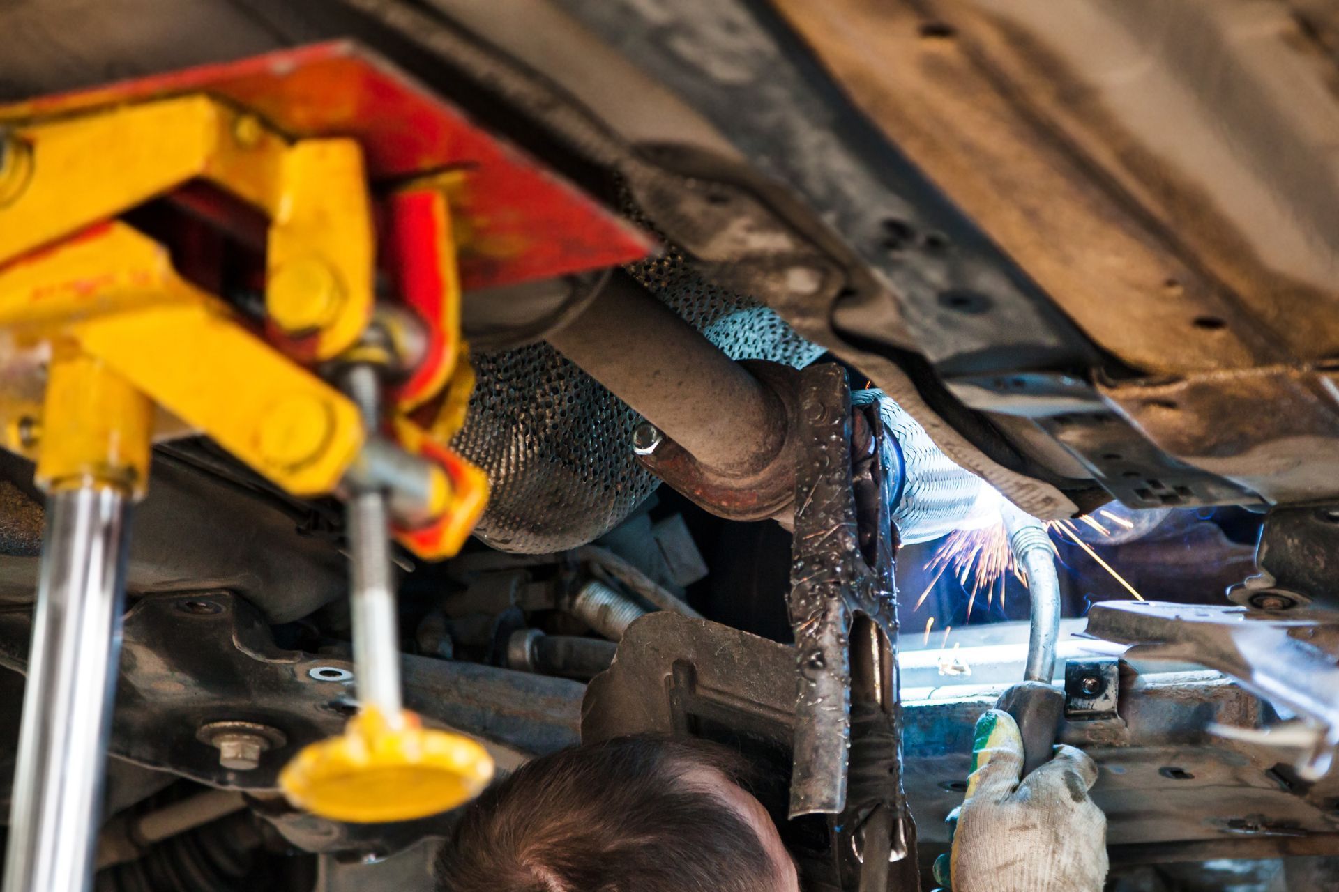 A man is welding under a car with a yellow jack.
