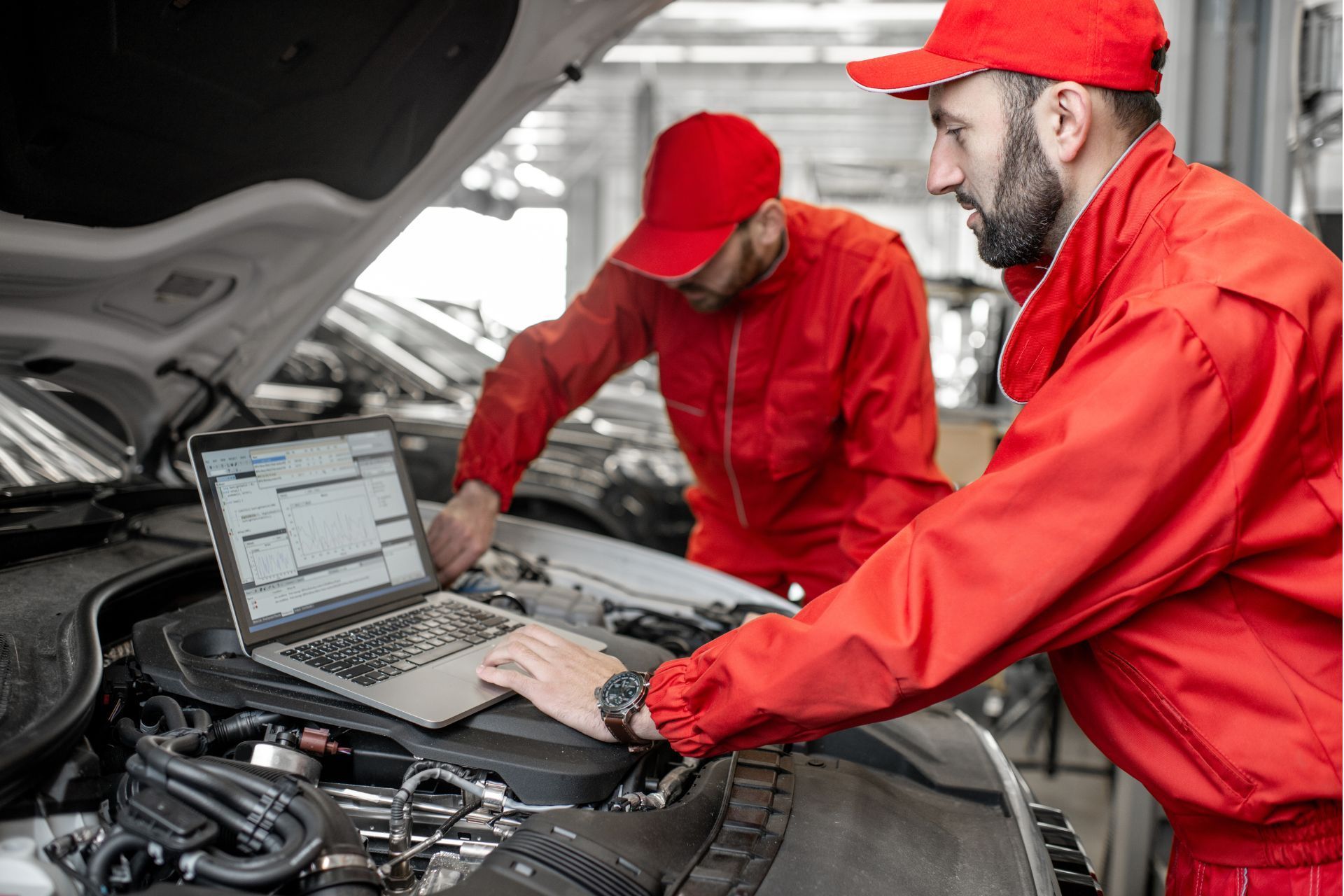 Two mechanics are working on a car engine with a laptop.
