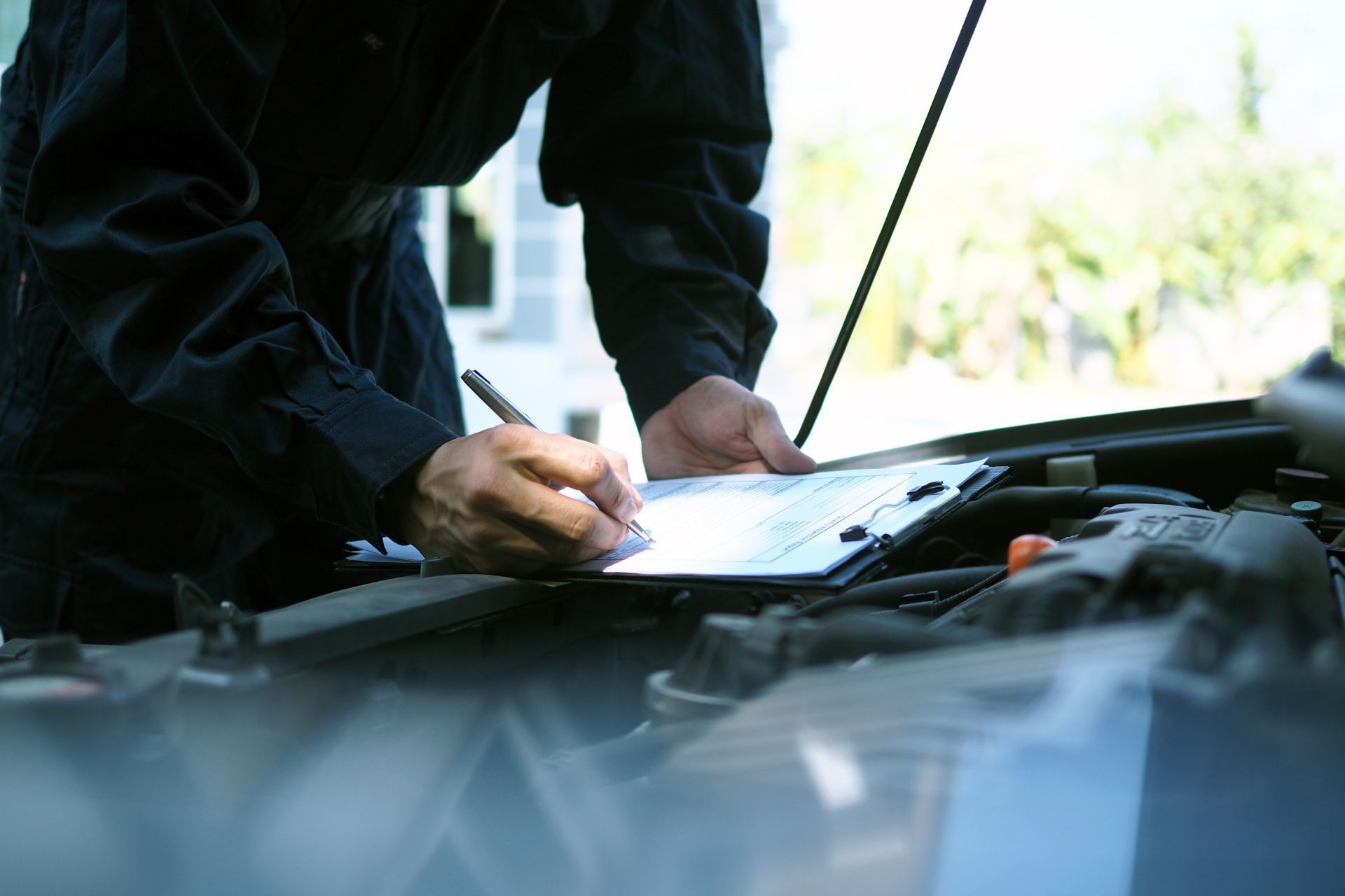 A man is writing on a clipboard under the hood of a car.