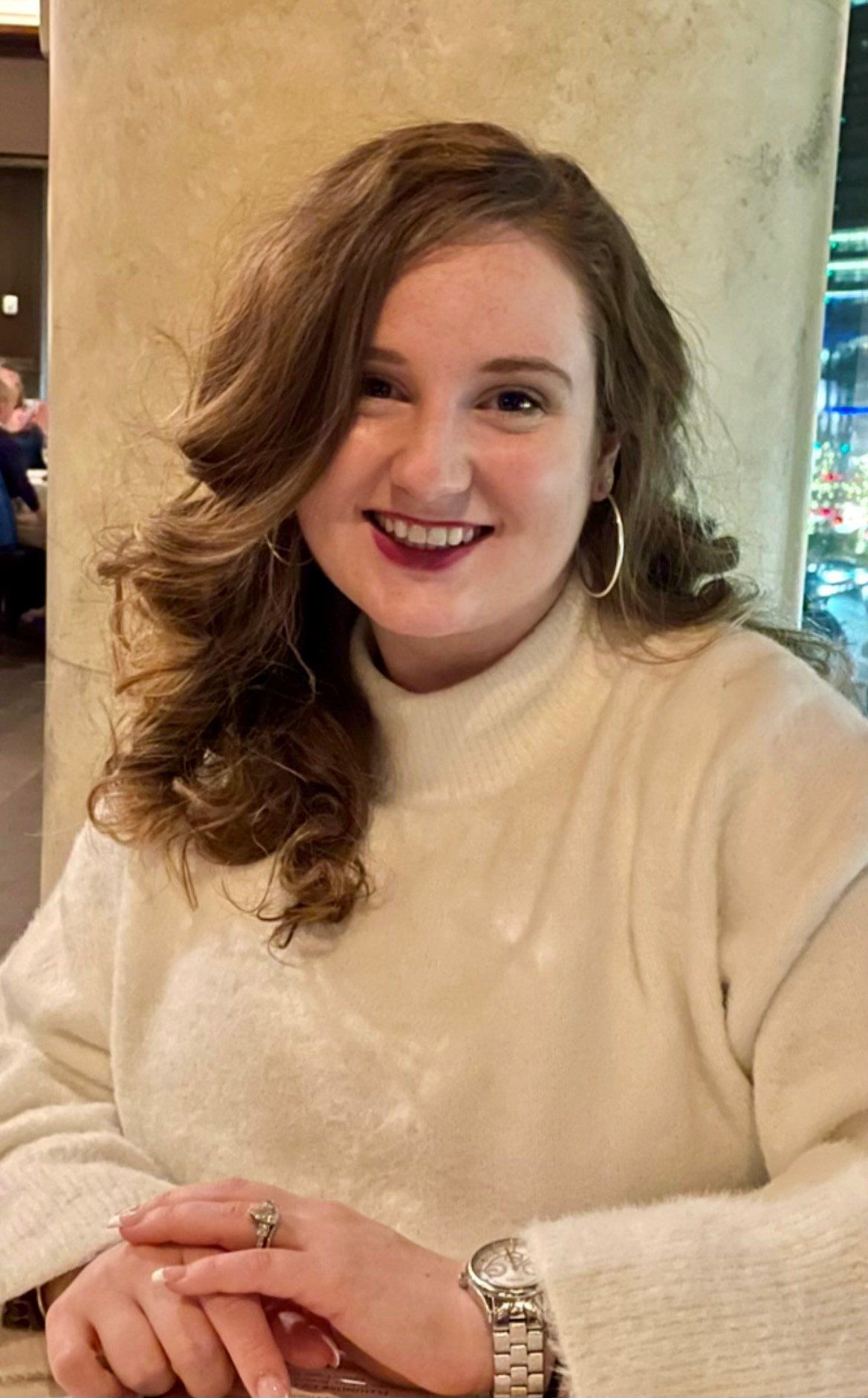 A woman wearing a white sweater and a watch is smiling while sitting at a table.