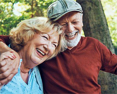 An elderly couple is holding hands and smiling while sitting under a tree.