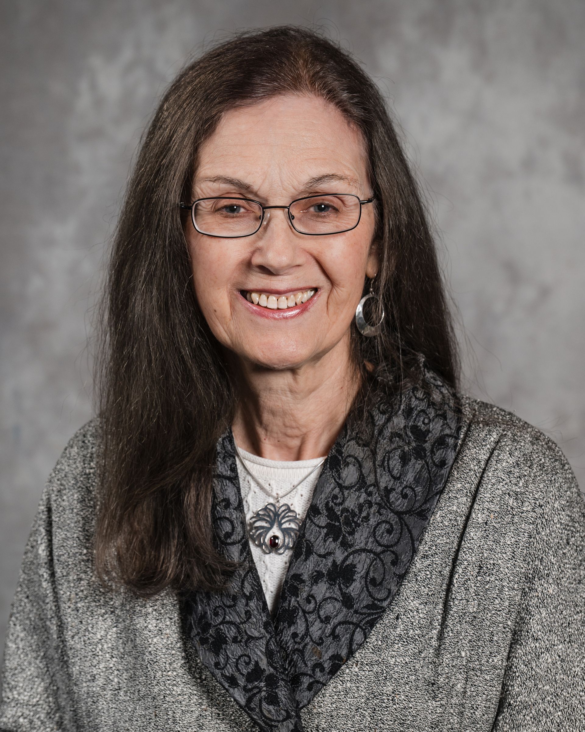 A smiling person wearing glasses, a grey textured jacket, and a dark patterned scarf against a grey background.