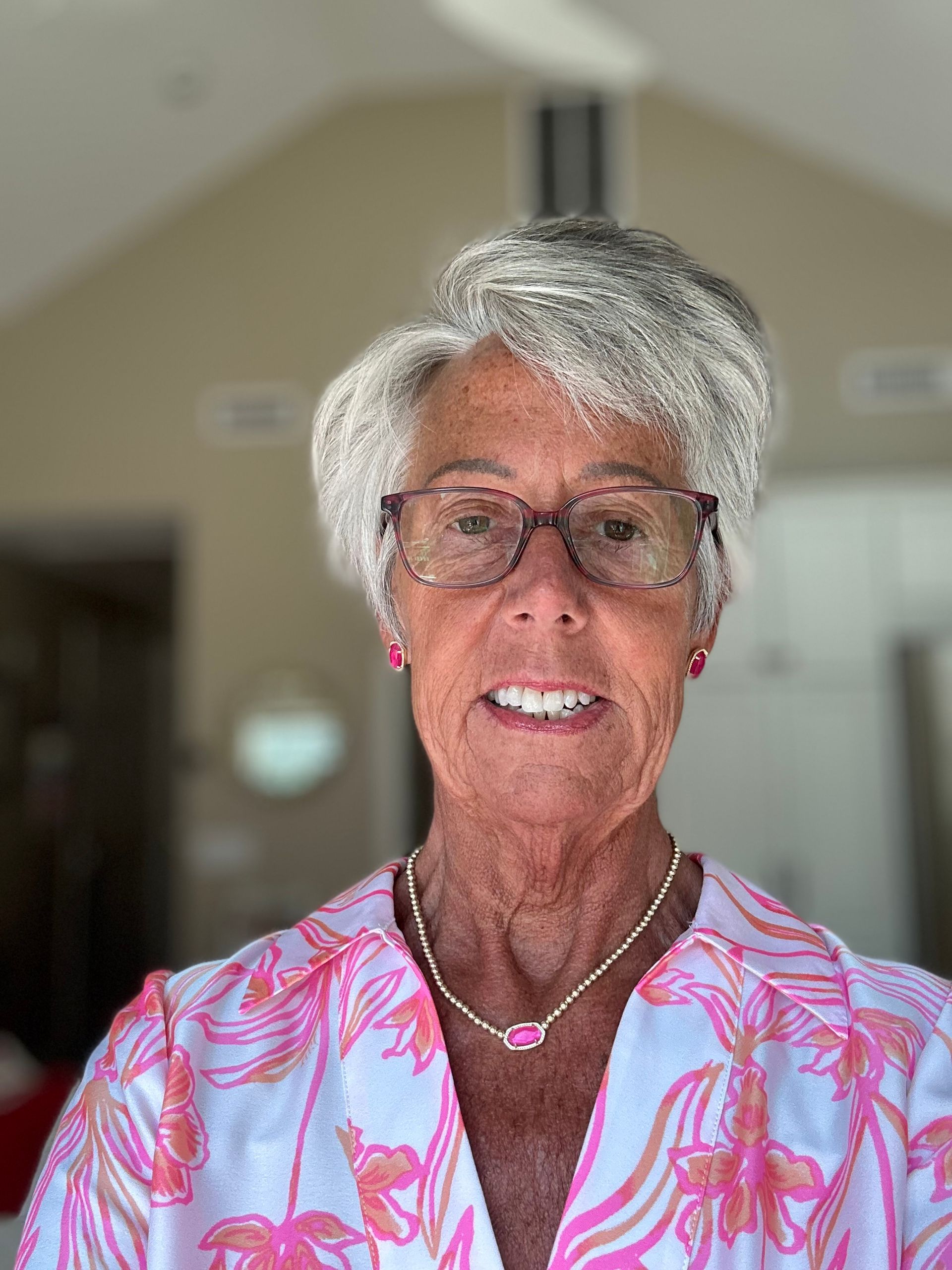 A person with short, silver hair and glasses wears a pink and white floral top and a necklace against a blurred background.
