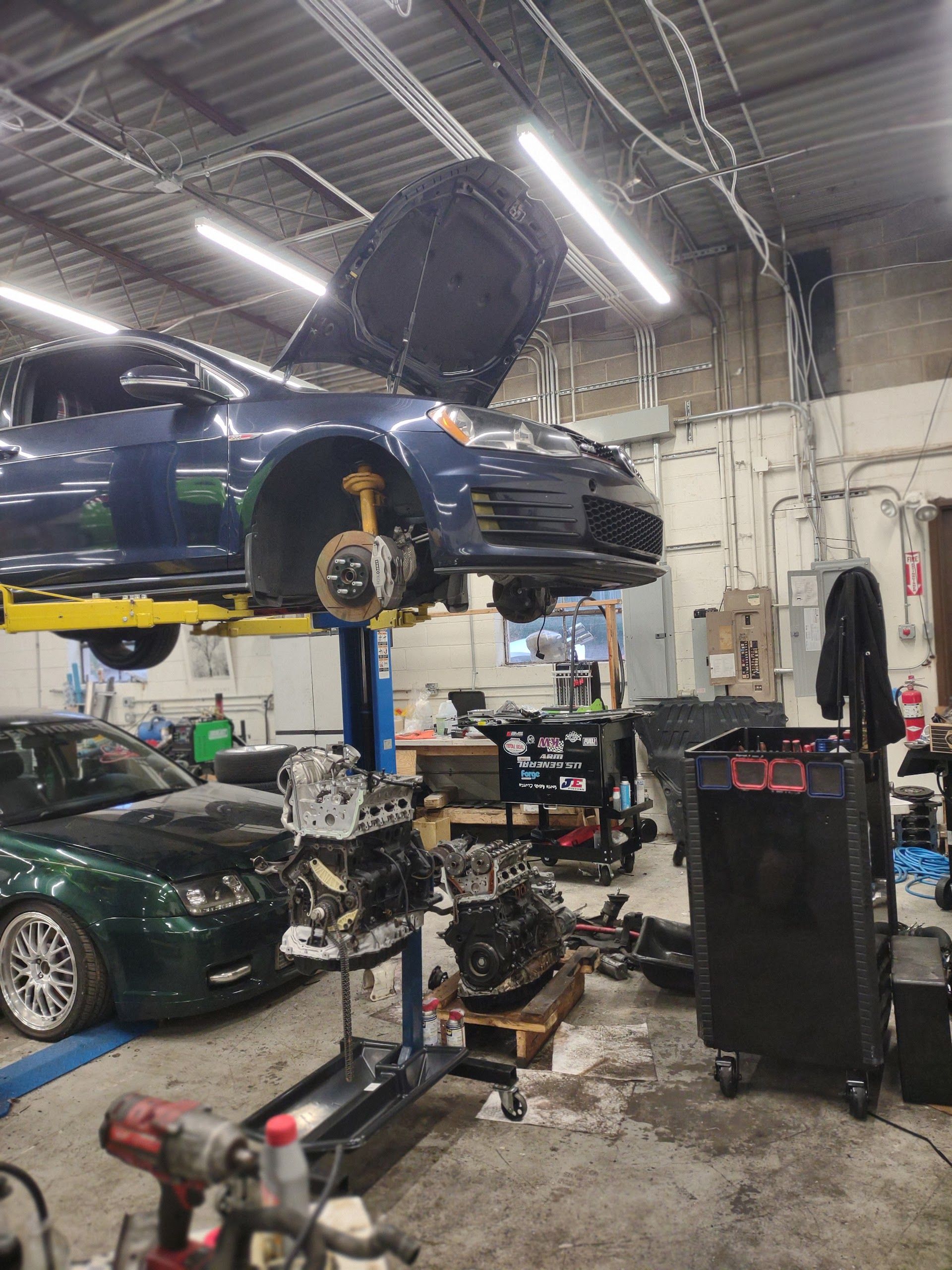 Car on a lift in a repair shop with hood open, other cars and engines visible. | A2B Euro Car Repair