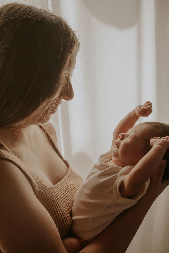 Woman holding a baby, looking down. Baby is stretching with eyes closed. Soft lighting, neutral tones.