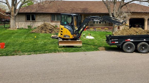 Yellow excavator on grass next to a black trailer, in front of a house.