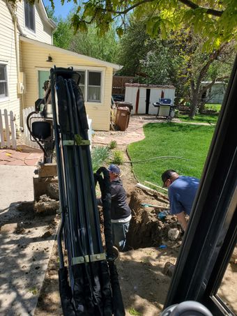 Workers dig a trench in a backyard. A small excavator is in the foreground. A house, shed, and grill are in the background.