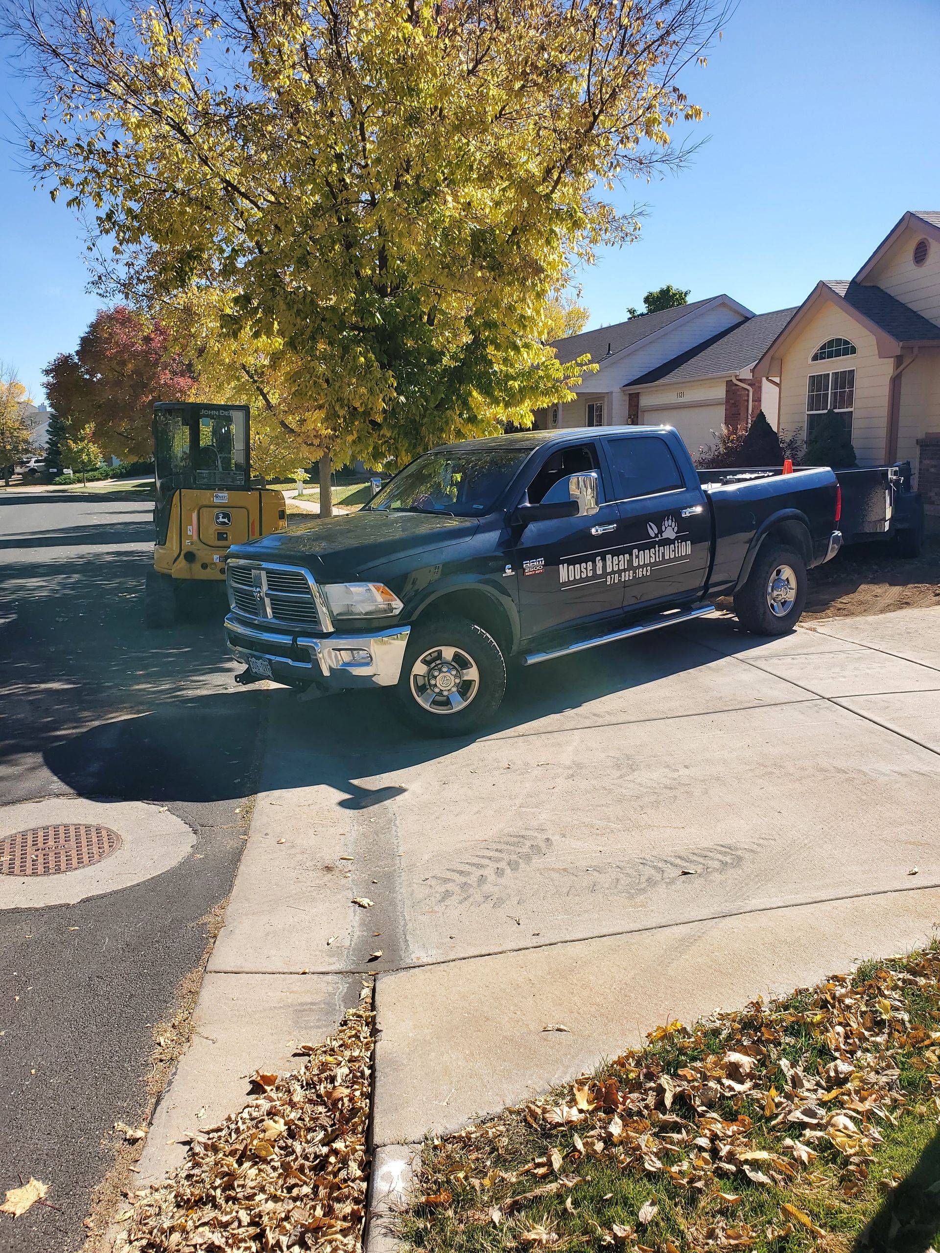 Black pickup truck and trailer parked on a driveway near a home, with a yellow construction vehicle in the background.