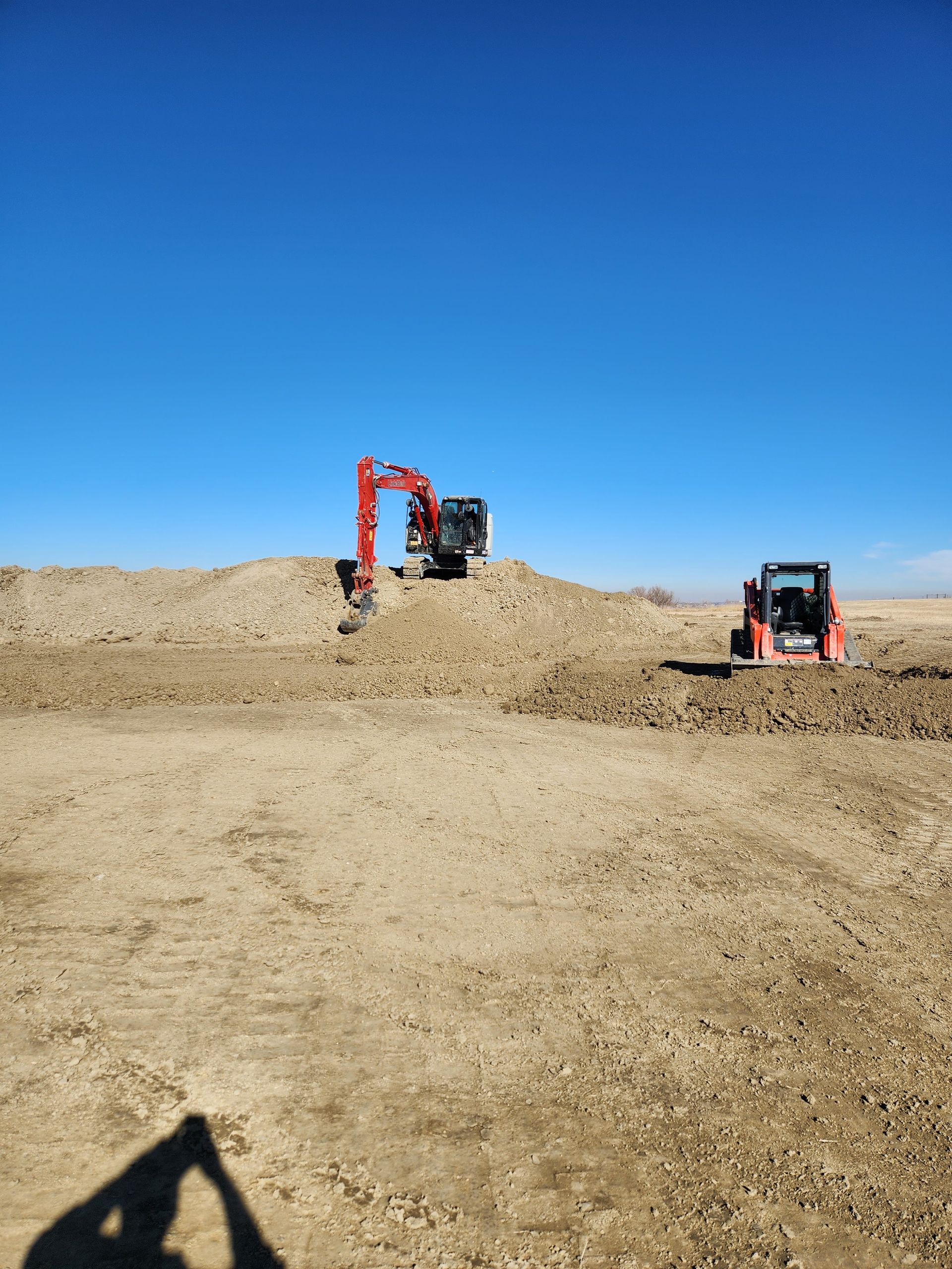 An excavator and skid steer on a dirt lot under a clear blue sky.