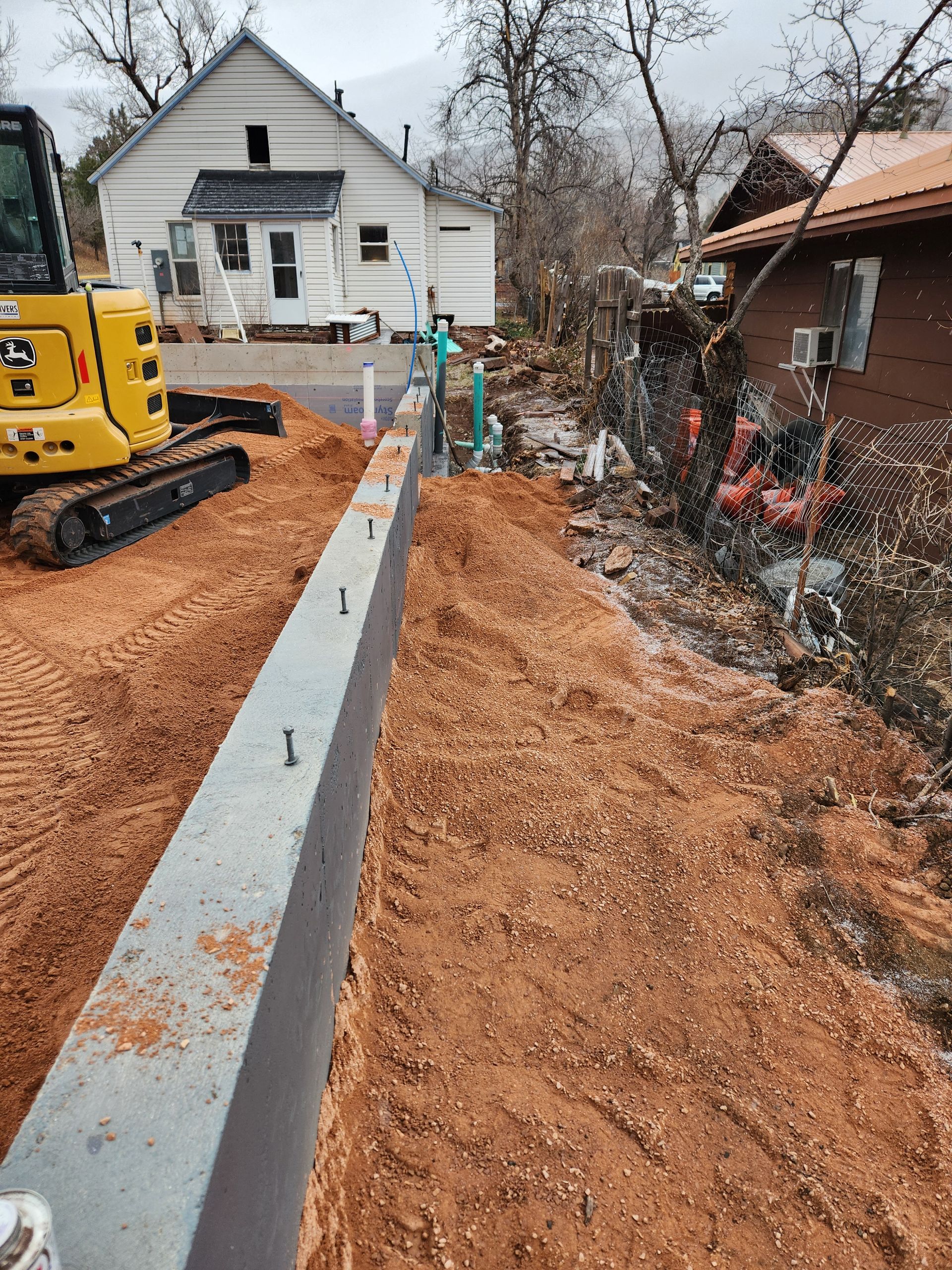Workers dig a trench in a backyard. A small excavator is in the foreground. A house, shed, and grill are in the background.