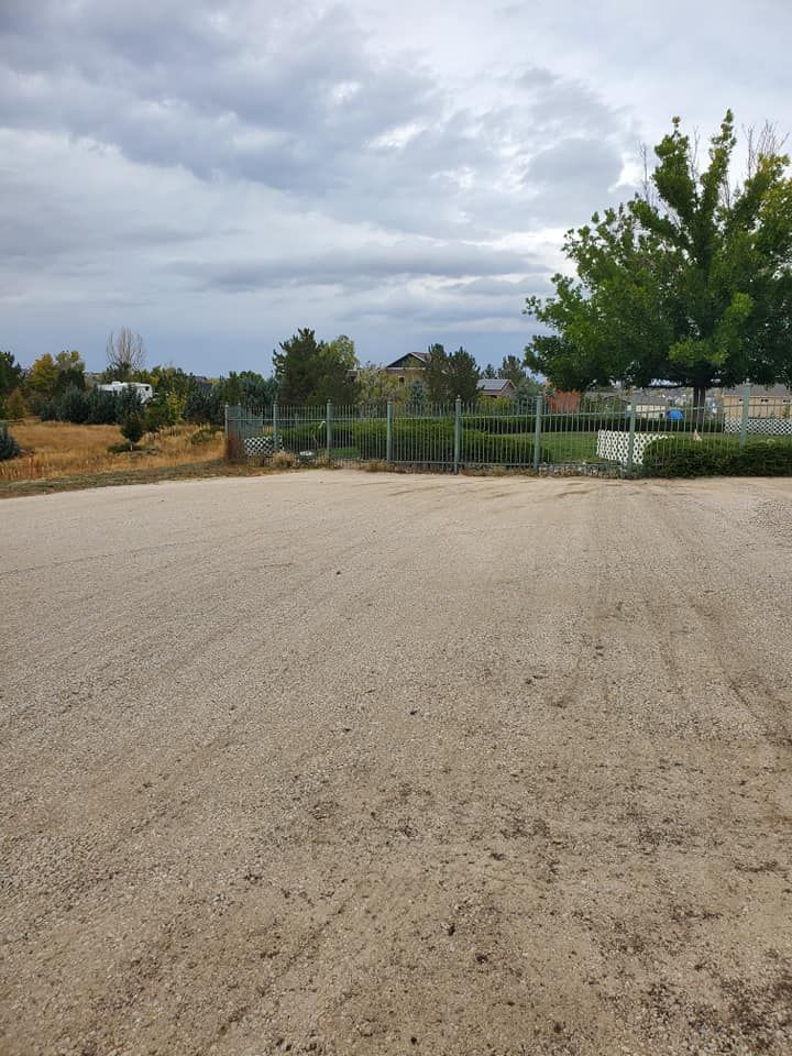 Gravel parking area leads to a fenced-in field with trees and buildings under a cloudy sky.