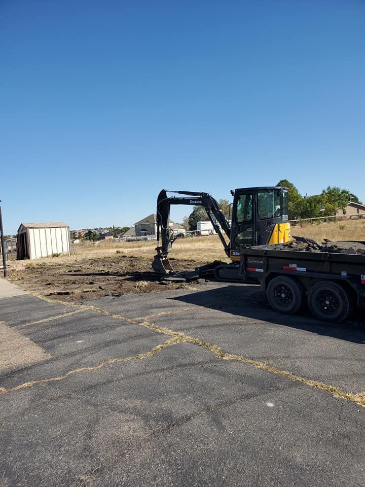Excavator on a brown dirt lot, trailer nearby, clear blue sky.