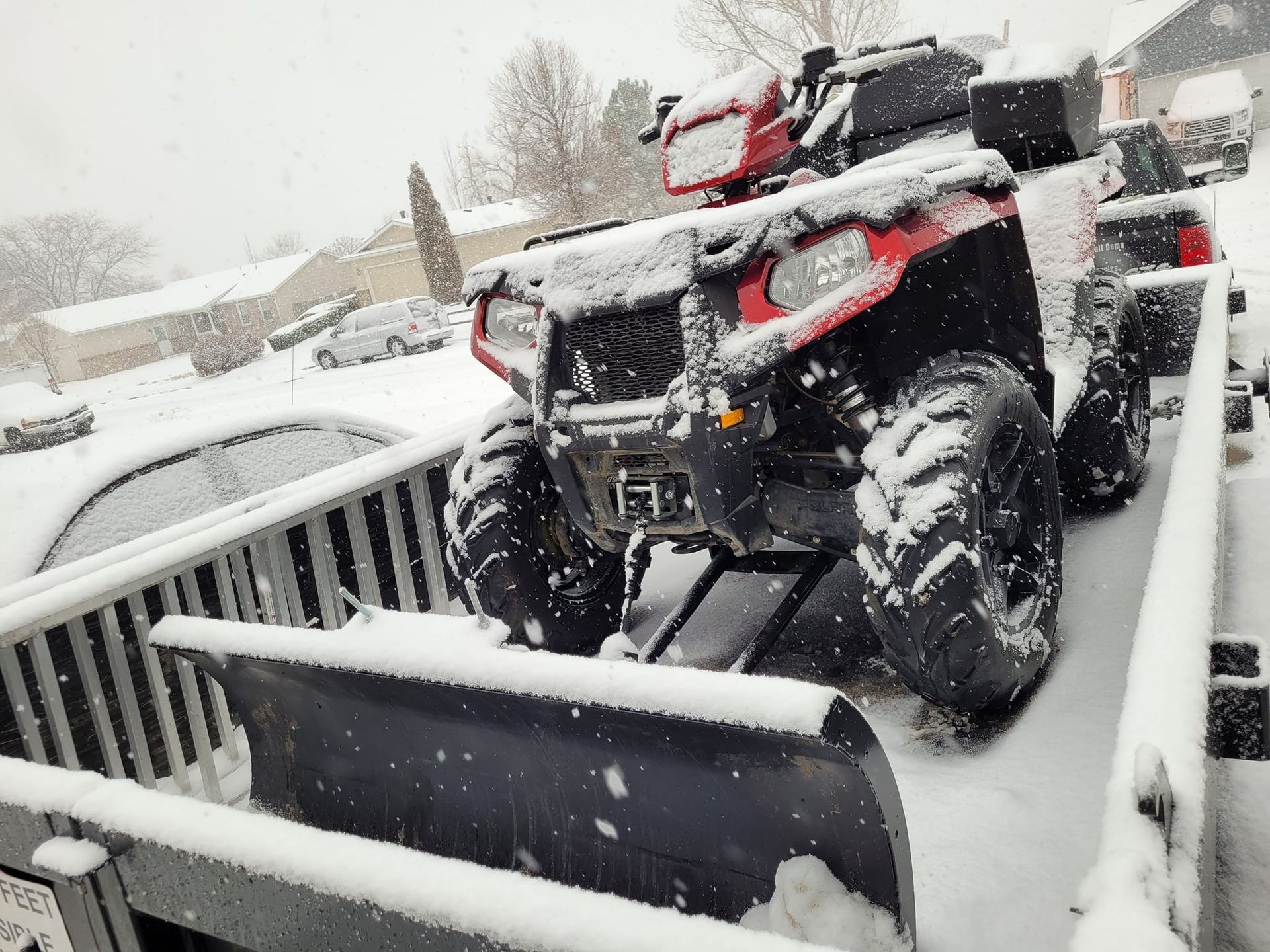 Red and black ATV with snow plow clearing snow on a ramp. Snow covers vehicle, setting is outdoors.