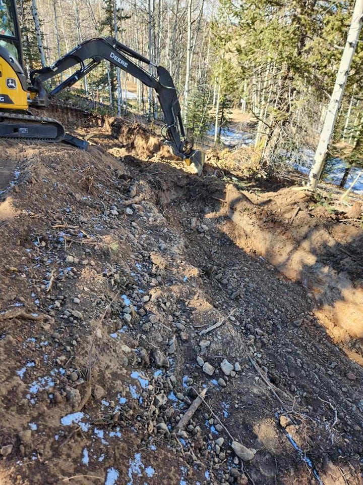 John Deere excavator digging into a hillside in a wooded area. Brown soil, some snow, and trees are visible.