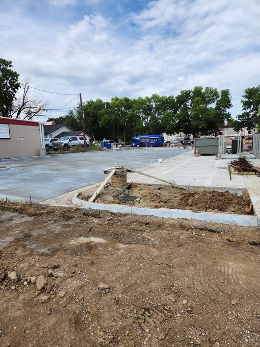 Construction site with dirt, gravel, and concrete. Buildings and trees in the background under a cloudy sky.