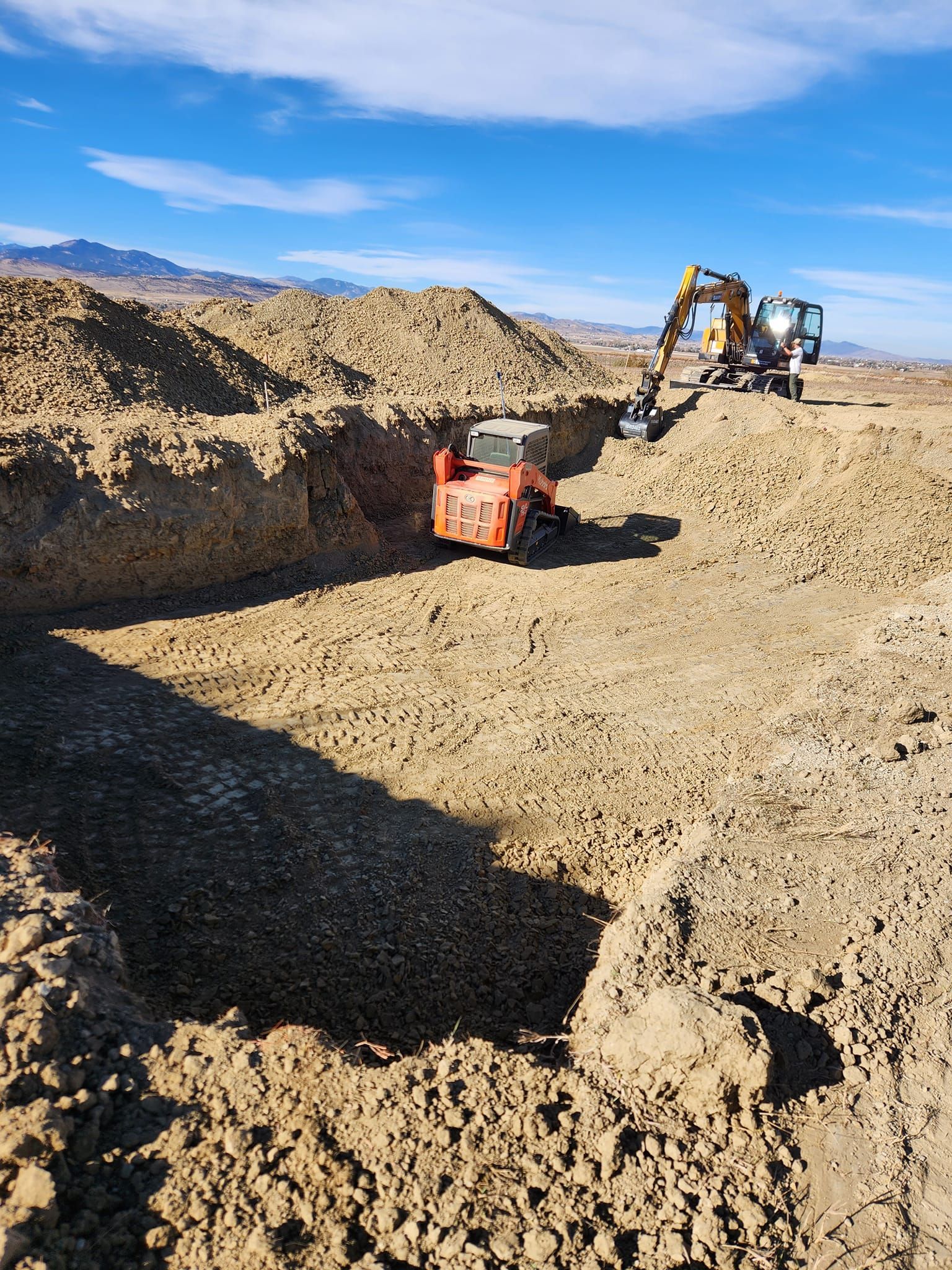 Two excavators digging a trench in a dirt field under a blue sky, mountains in the distance.
