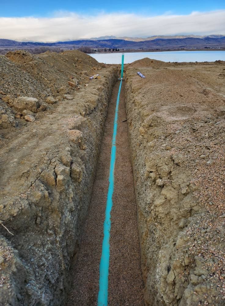 Blue pipe in a trench with gravel, in an outdoor setting with a body of water and hills in the background.