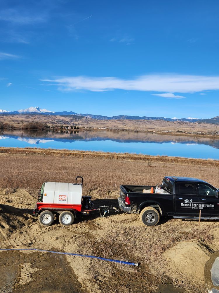 Black truck towing a trailer with a tank, near a lake with mountains in the background under a blue sky.
