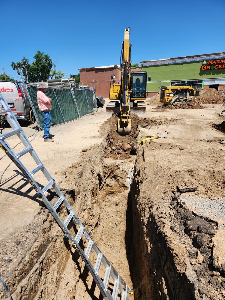 An excavator digs a trench in a construction site. A man watches, a ladder leans inside the trench. Blue sky.