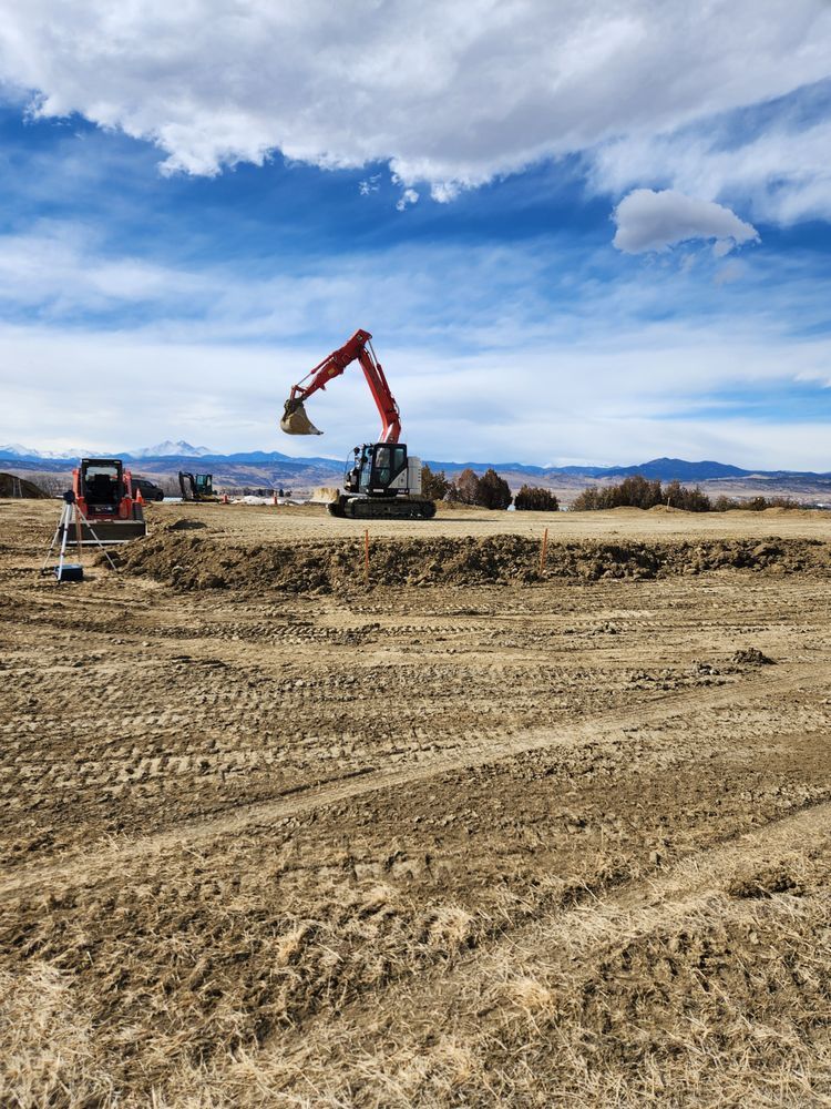 Excavator digging in dirt field under cloudy blue sky. Another small excavator sits nearby.