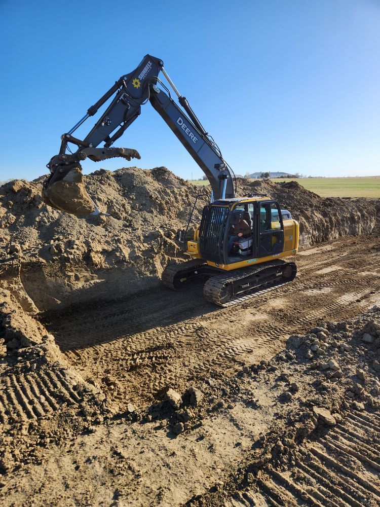 Excavator digging a trench in a sandy area under a clear blue sky.