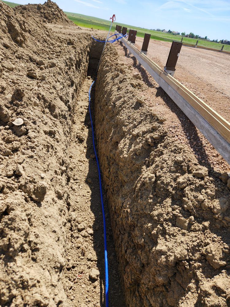 Trench dug in dirt, blue pipe inside. Wooden beam and fence posts along the side, road, sky, and flag in the background.