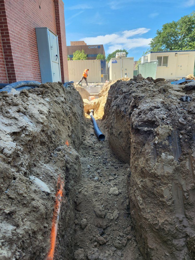 Construction site with a trench, pipe, and worker walking. Brick building on the left.