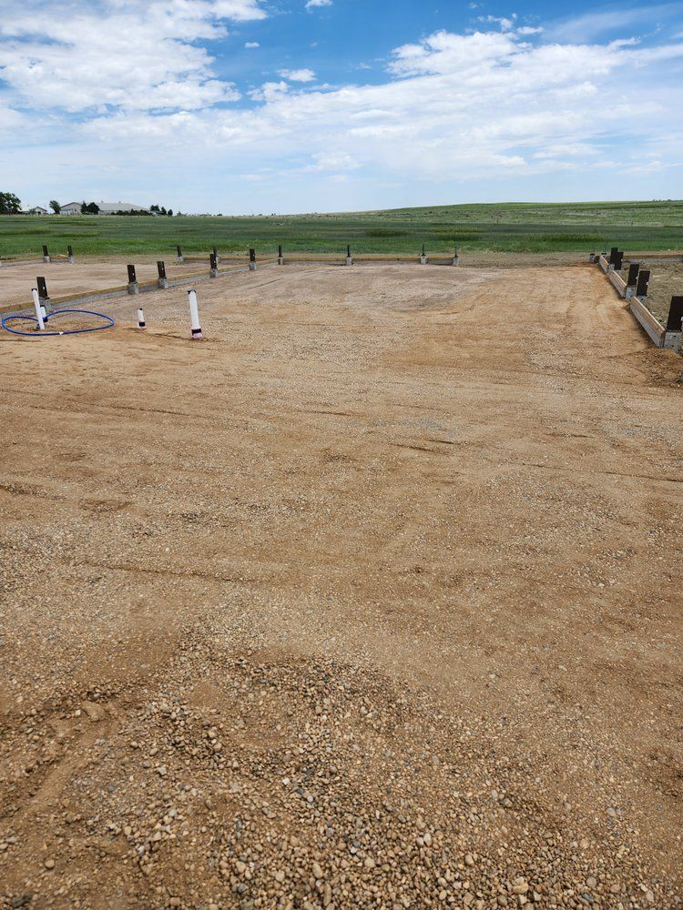 A gravel RV pad with utility hookups, poles and grass field under a cloudy blue sky.
