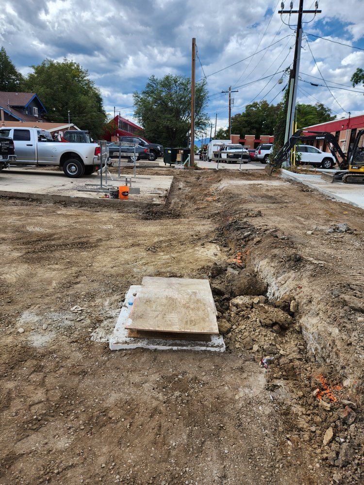 Construction site: trench, open manhole cover, dirt, vehicles and buildings in background.