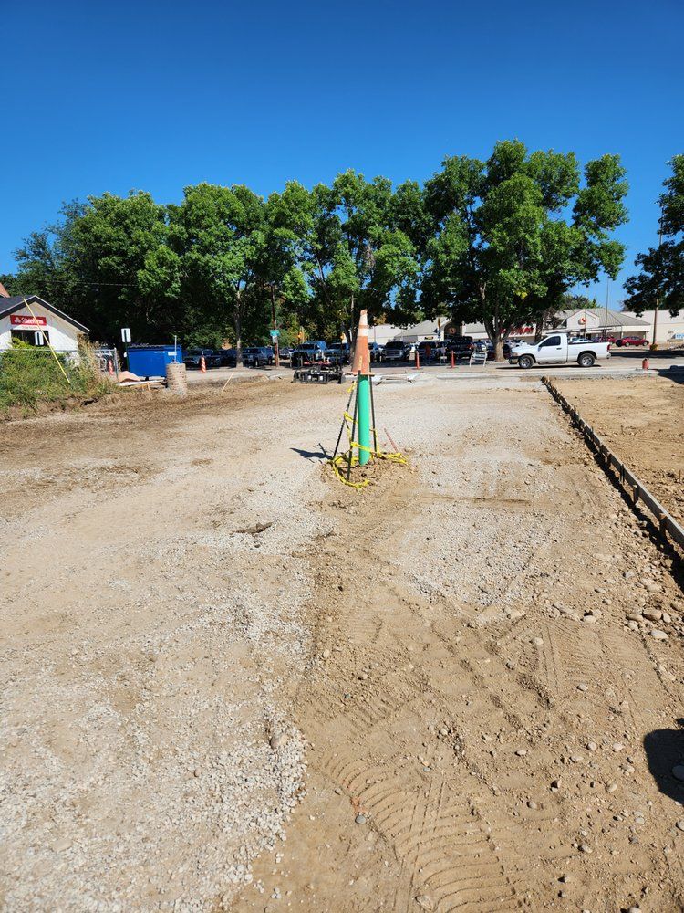 Construction site with gravel ground and a green pipe. Vehicles and trees are in the background.