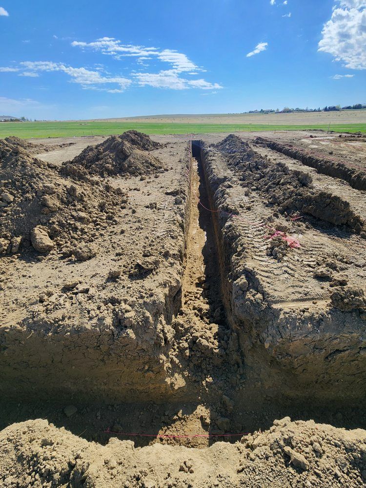 Long trench dug in a field under a blue sky, dirt piles on either side, with distant farmland.