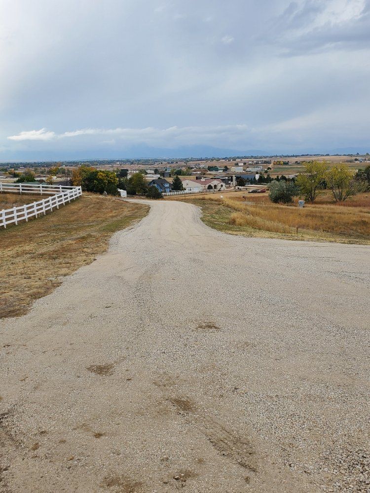 Dirt road leading to a suburban neighborhood under a cloudy sky. White fence on left, dry field.
