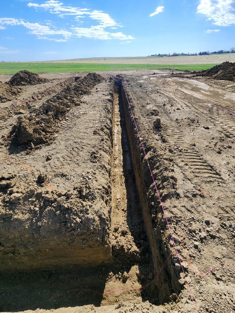 A long, narrow trench dug in brown soil, leading toward a green field under a blue sky.