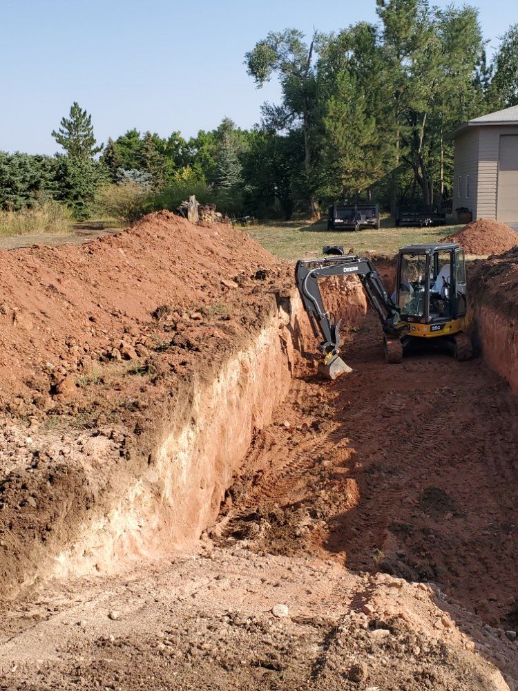 Mini excavator digging a long trench in red soil, near trees.