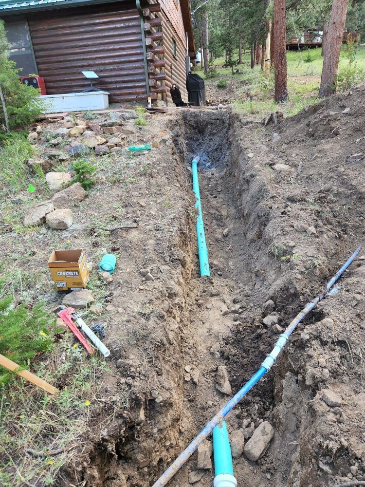 Trench dug with blue pipes running through it, beside a log cabin and trees.
