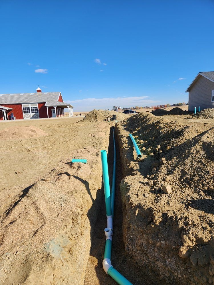 Trench with turquoise PVC pipes installed. Dirt mounds, buildings, and blue sky.