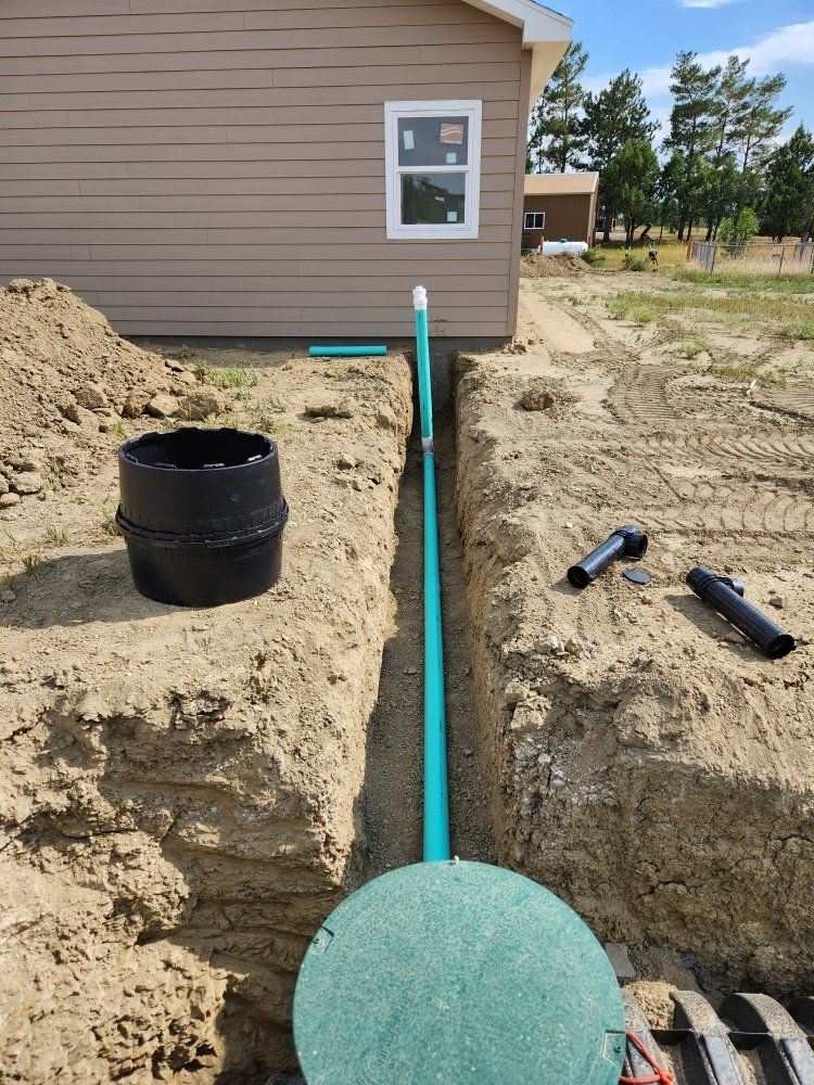 Construction site with trench, green pipe, and septic tank lid near a tan building.