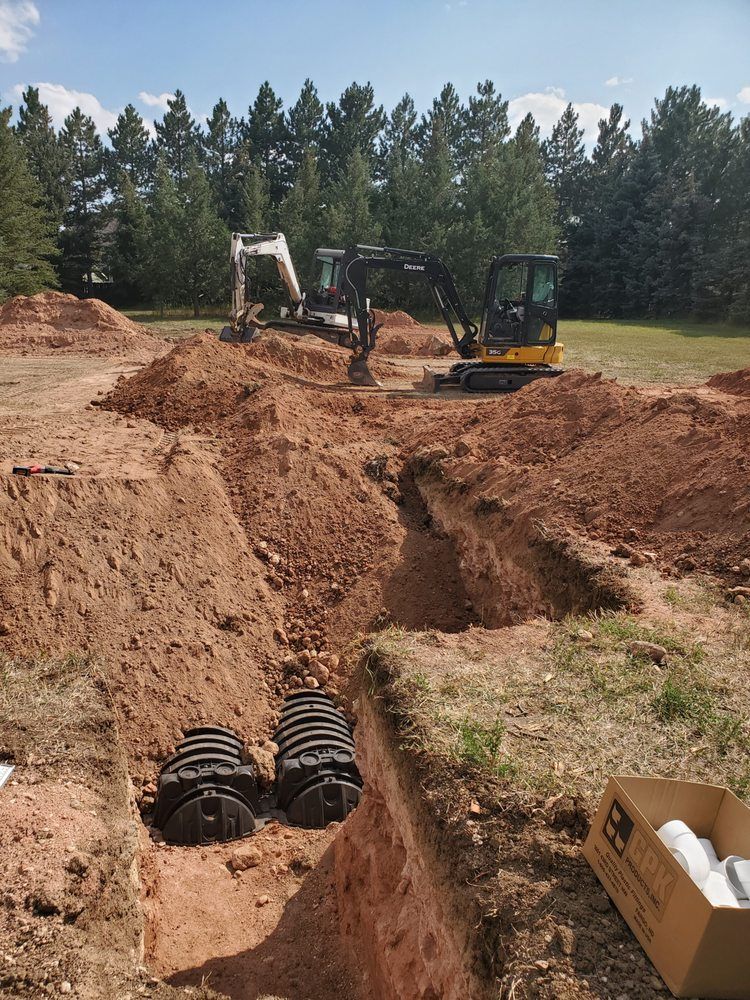 Excavators digging trenches in a dirt field for septic tank installation; trees in the background.