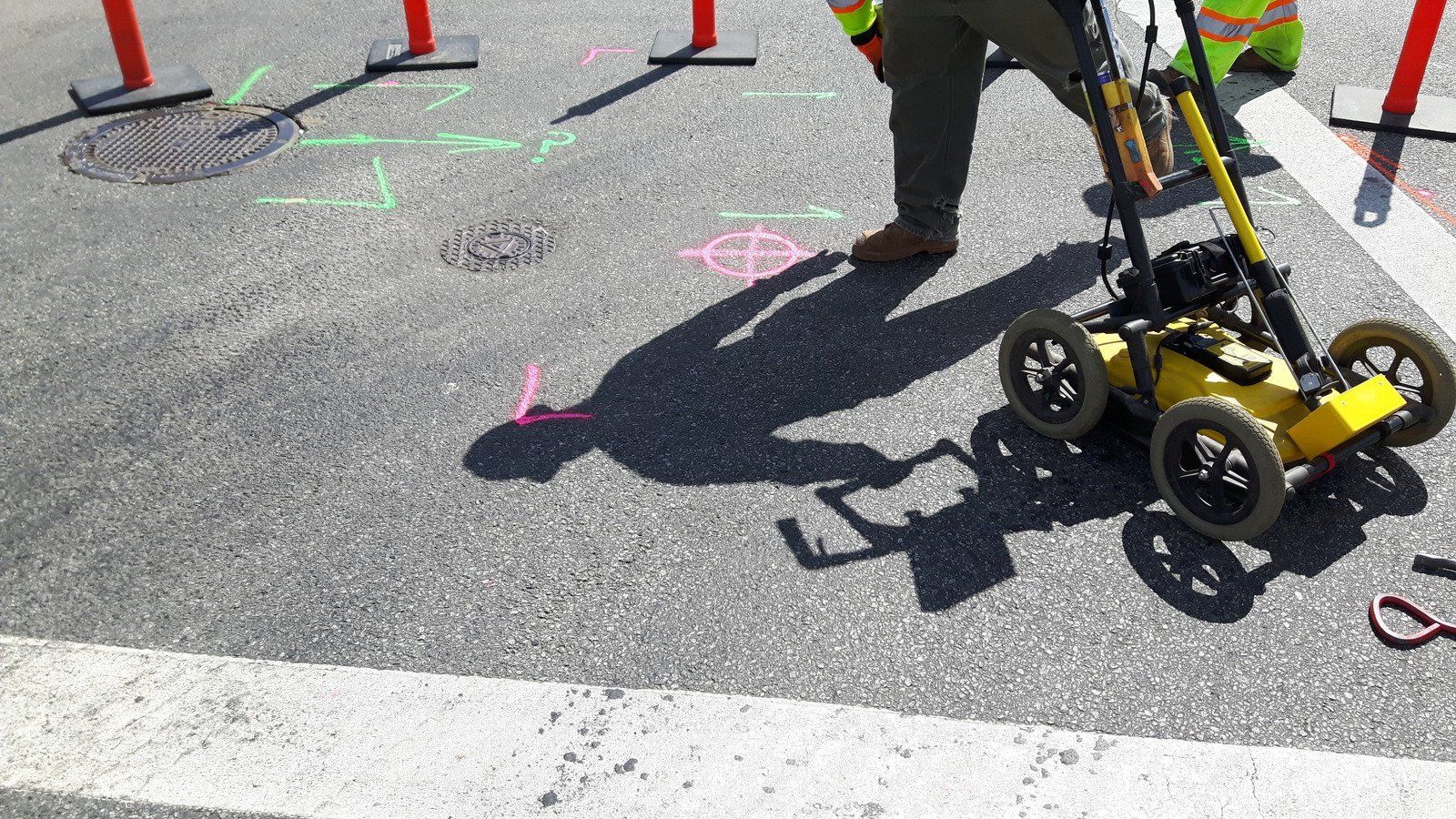 An excavator digs in a dirt lot in a forest, under a bright blue sky.