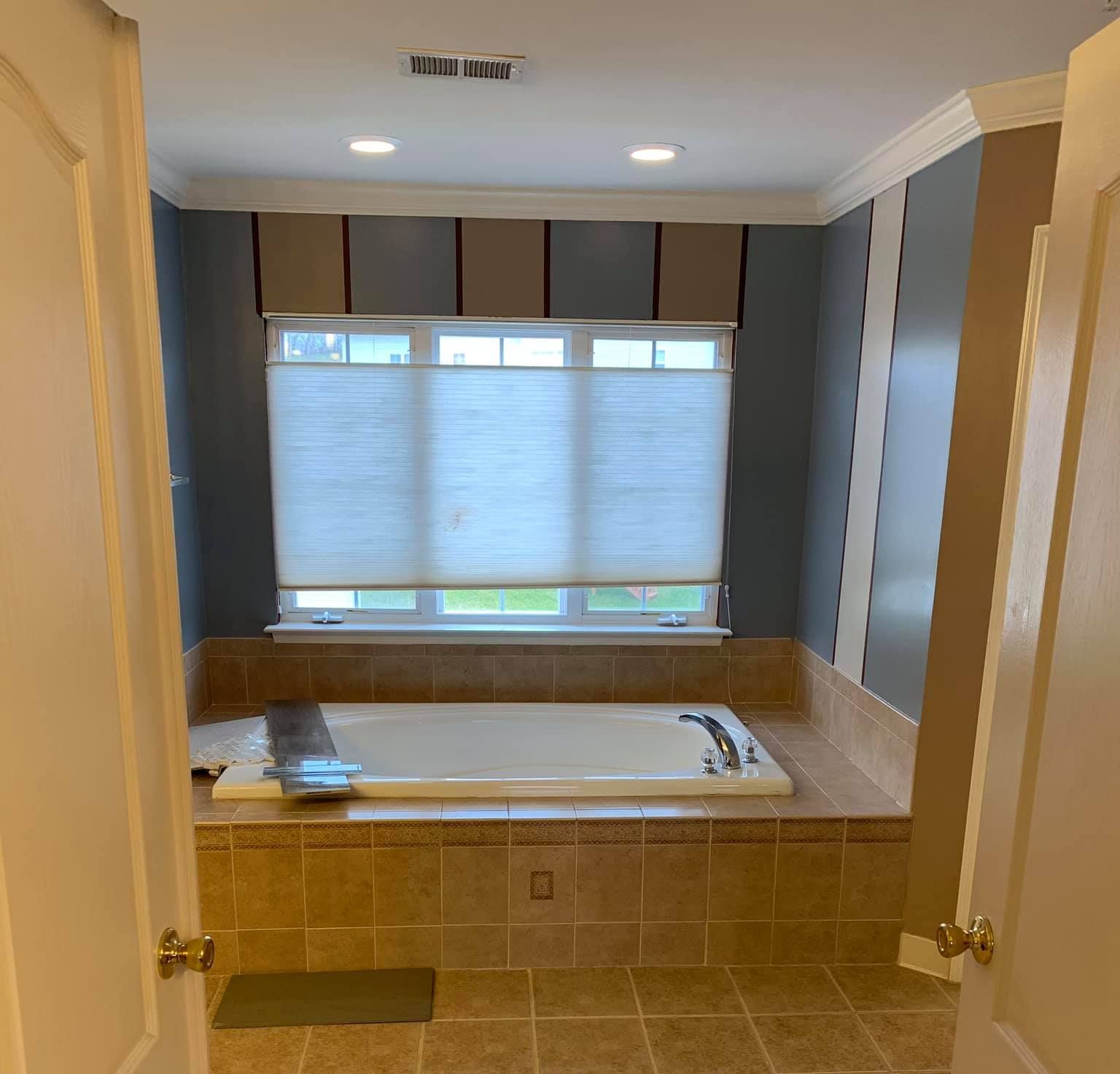 Bathroom with a built-in bathtub under a window. Beige and blue wall stripes, light-colored tile.