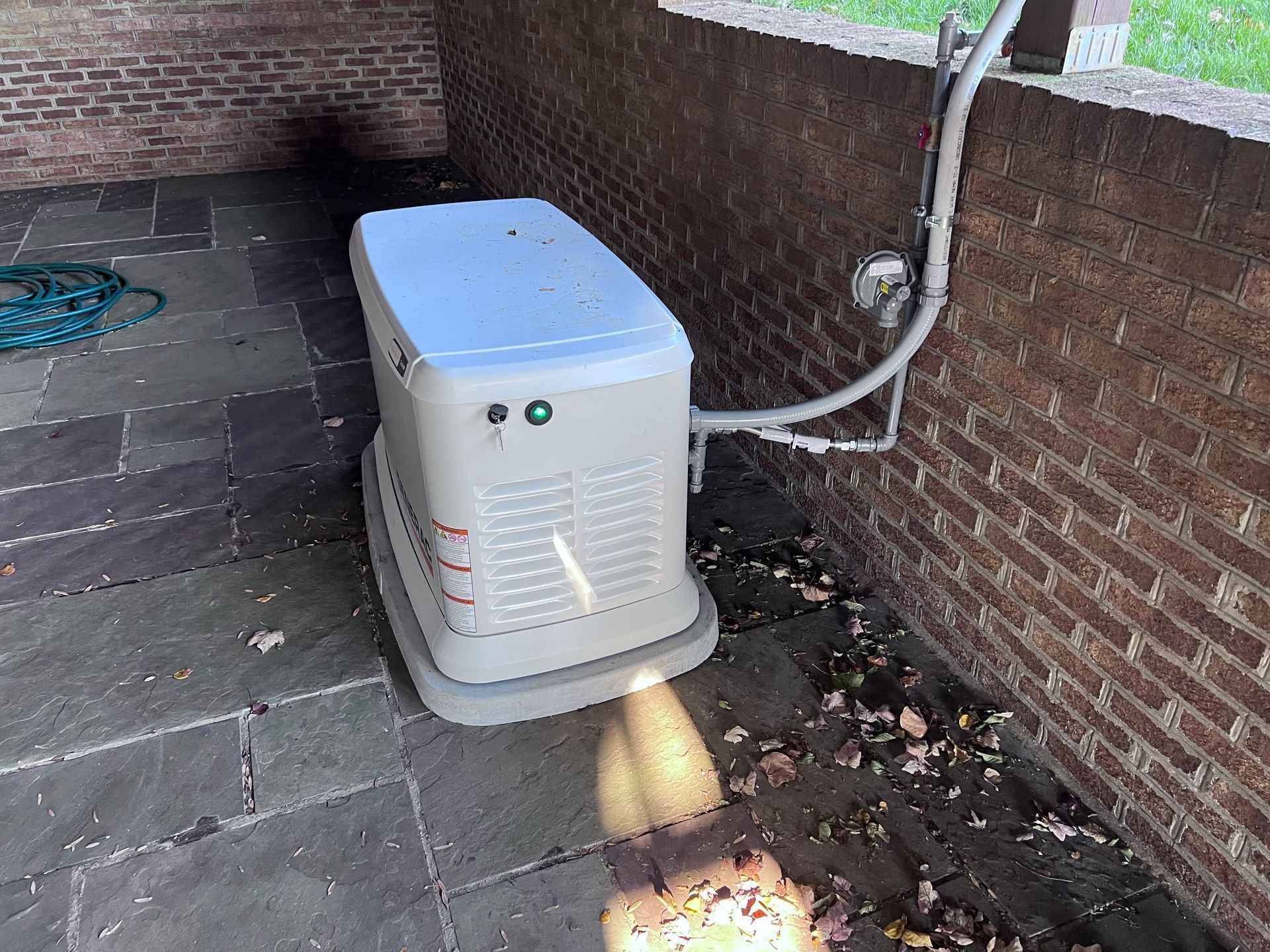 A tan standby generator sits on a stone patio against a brick wall, connected to the home by an exterior conduit pipe.