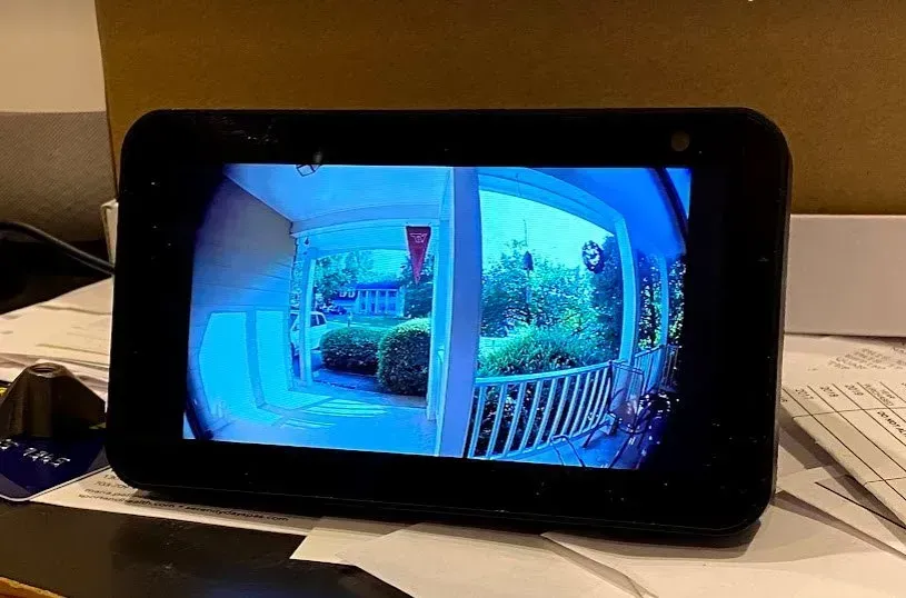 An Amazon Echo Show displays a wide-angle, blue-tinted video feed from a front porch camera showing greenery and a walkway.