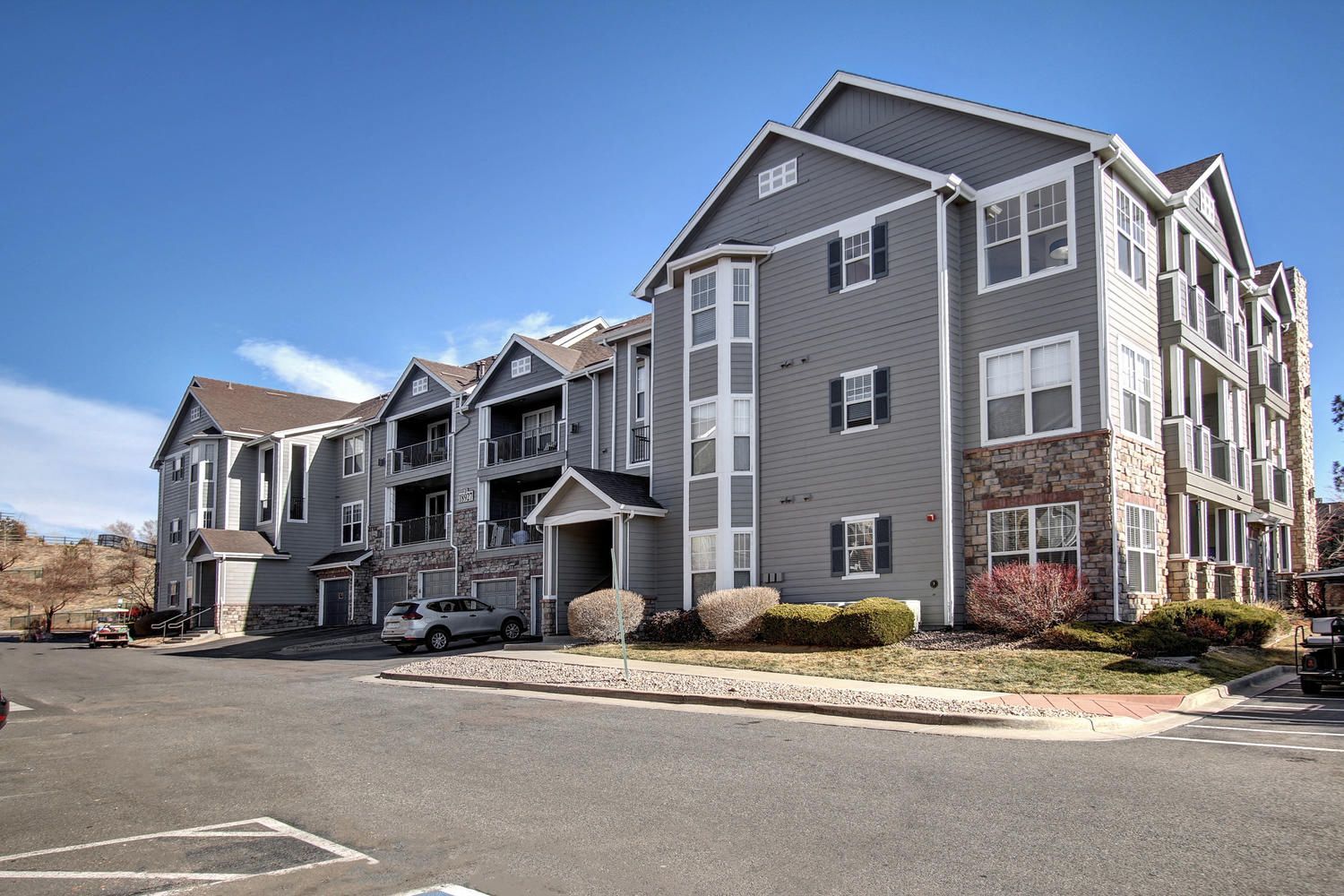Exterior view of a multi-building apartment complex with gray siding, stone accents, and a parking lot.