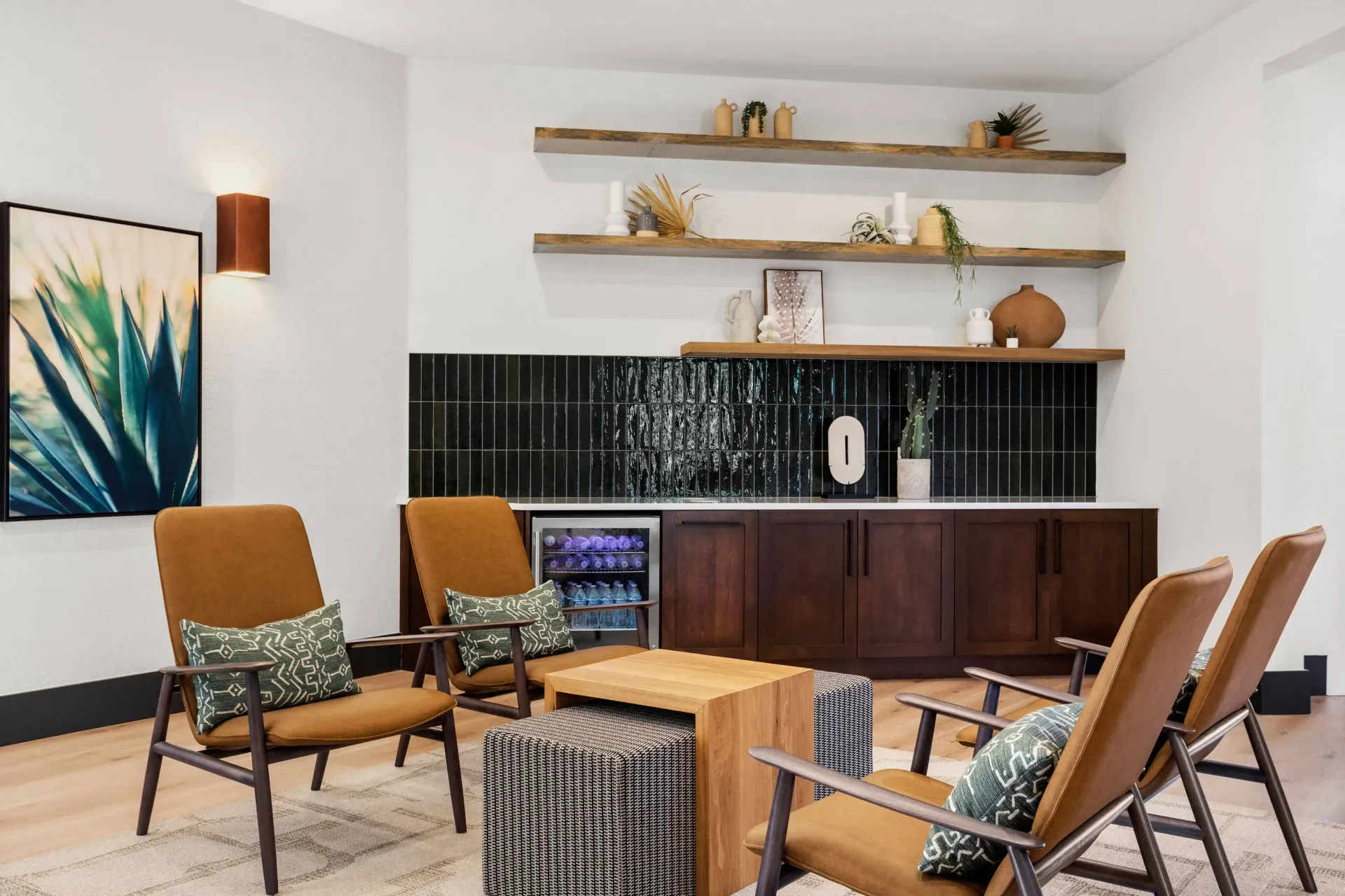 Communal lounge with tan mid-century chairs around a wooden coffee table and a dark tiled kitchenette.