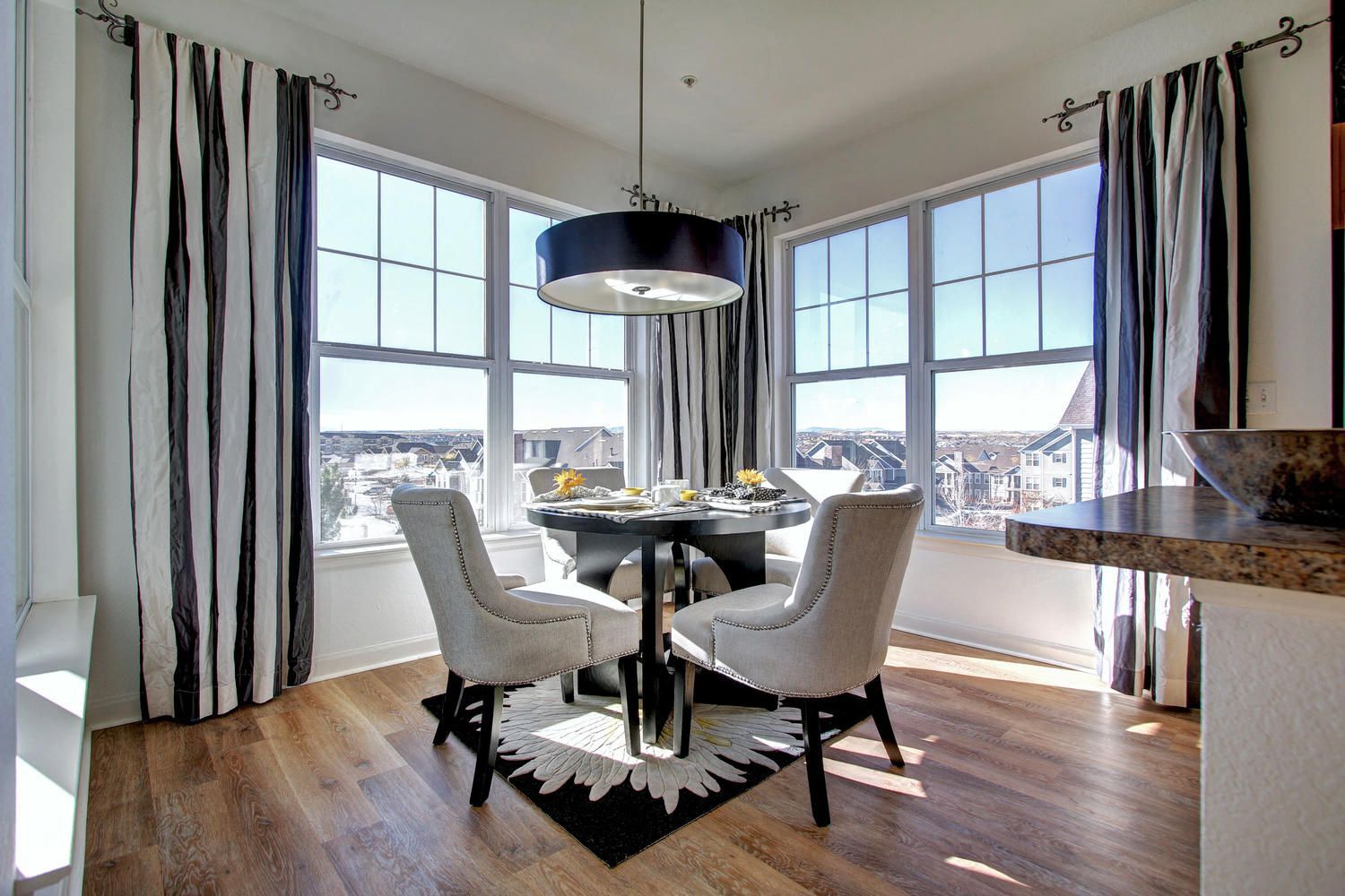 Round dining table with four upholstered chairs by large bay windows, sunlit.